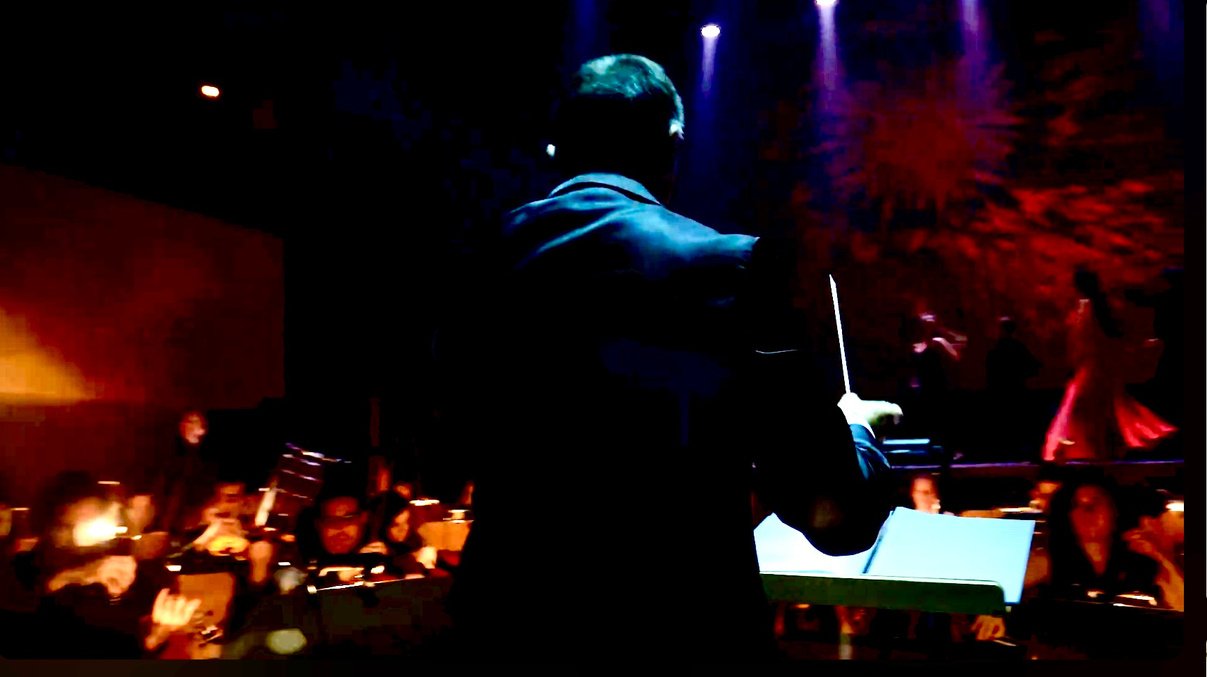 Conductor seen from behind leading an orchestra under dramatic lighting